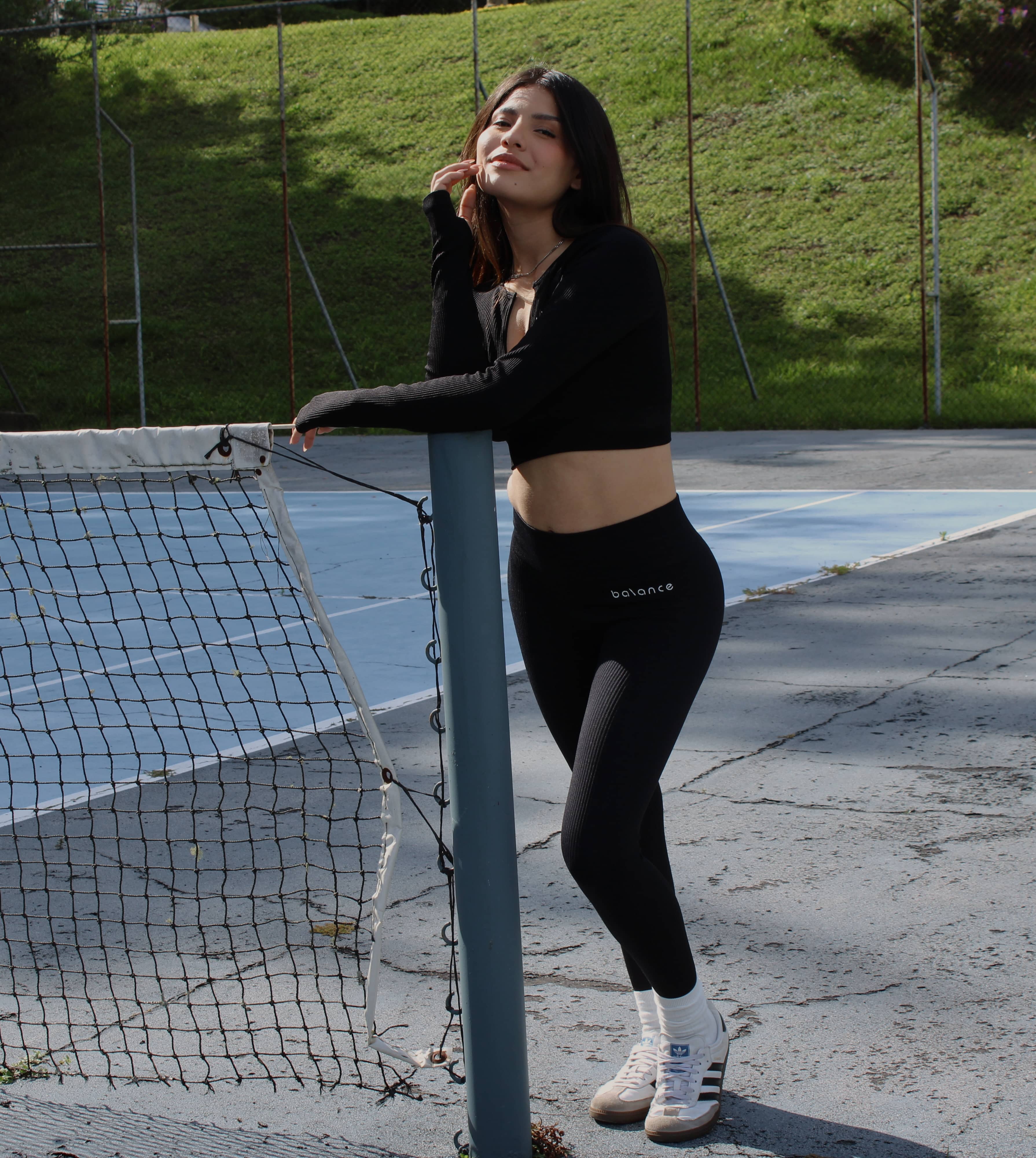 Modelo femenina de pie en una cancha deportiva al aire libre, usando un set de activewear negro con top de manga larga y legging de tiro alto.