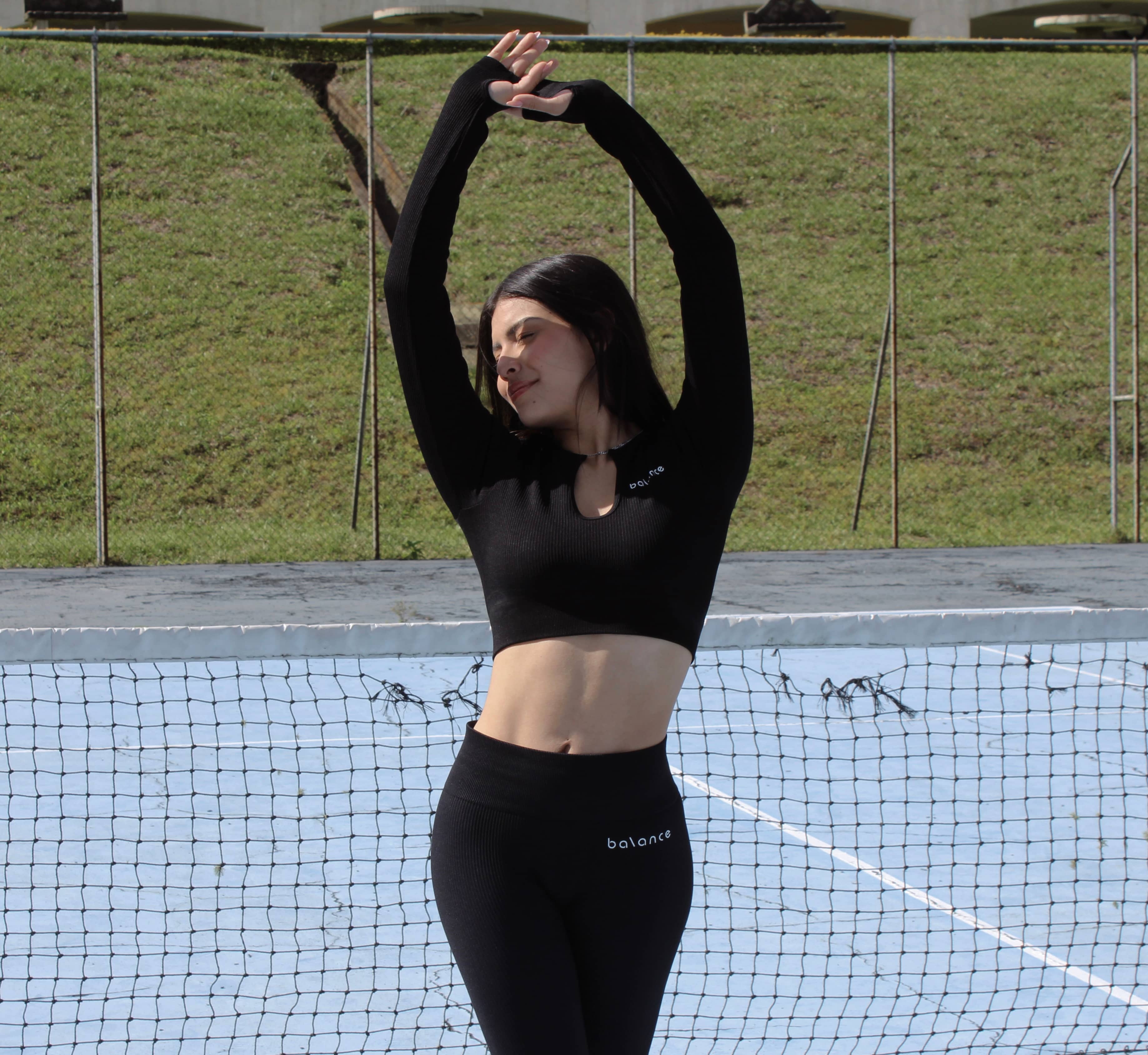 Modelo femenina estirando los brazos en una cancha deportiva, vistiendo un set de activewear negro con top de manga larga y legging de tiro alto.