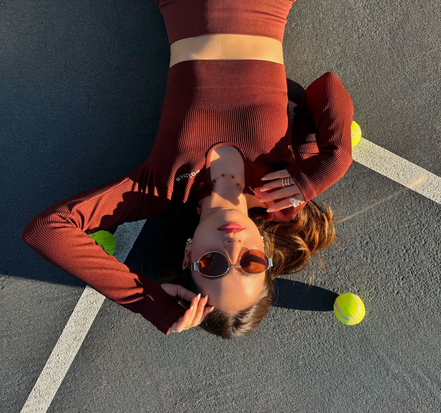 Modelo recostada en cancha con set deportivo color café de Balance y pelotas de tenis alrededor.