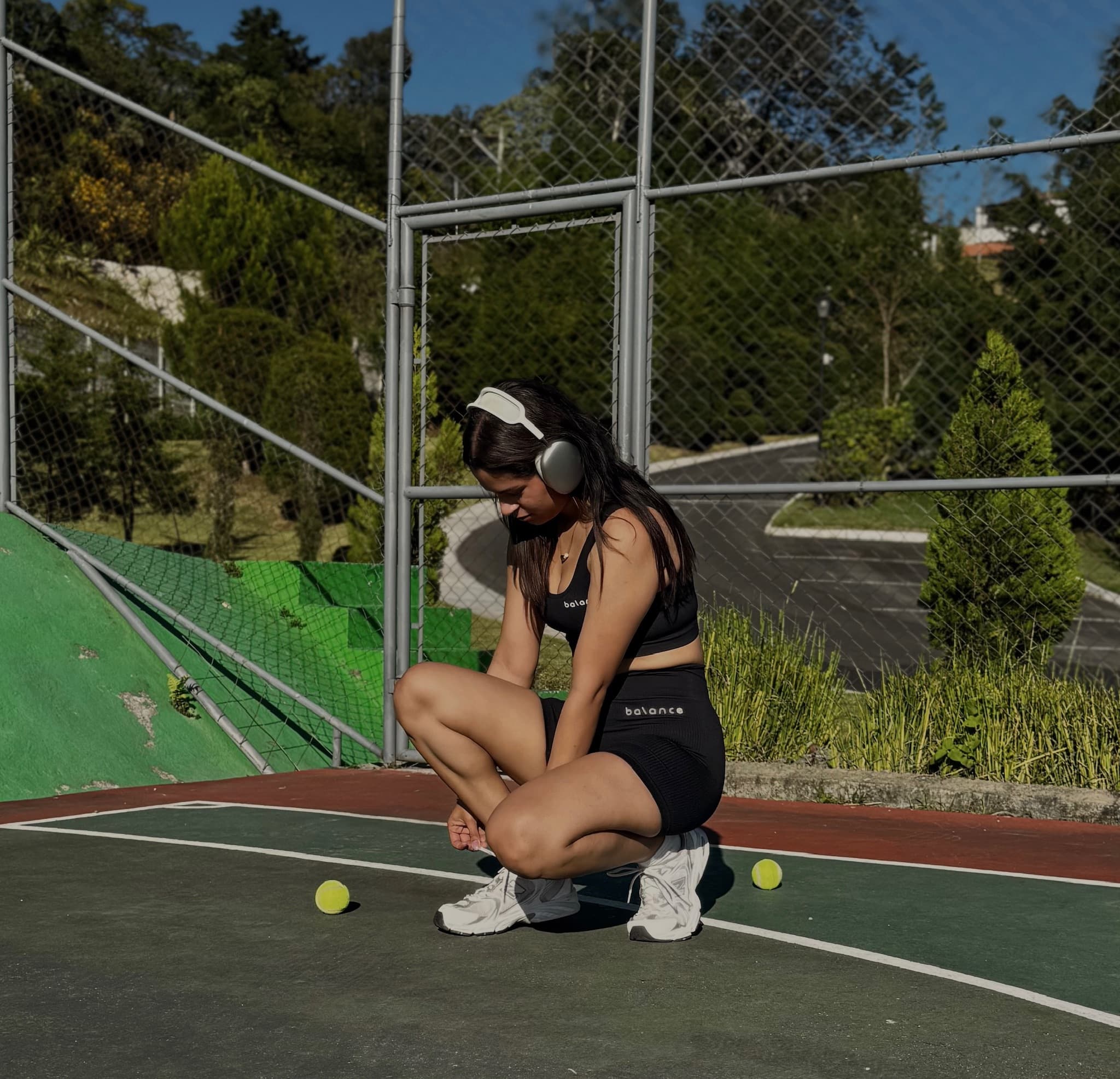Modelo femenina sentada en una cancha deportiva al aire libre, usando un set de activewear negro compuesto por un top sin mangas y un short de tiro alto con textura acanalada con el logo “Balance” visible en el top y el short. La escena transmite un estilo deportivo y relajado.