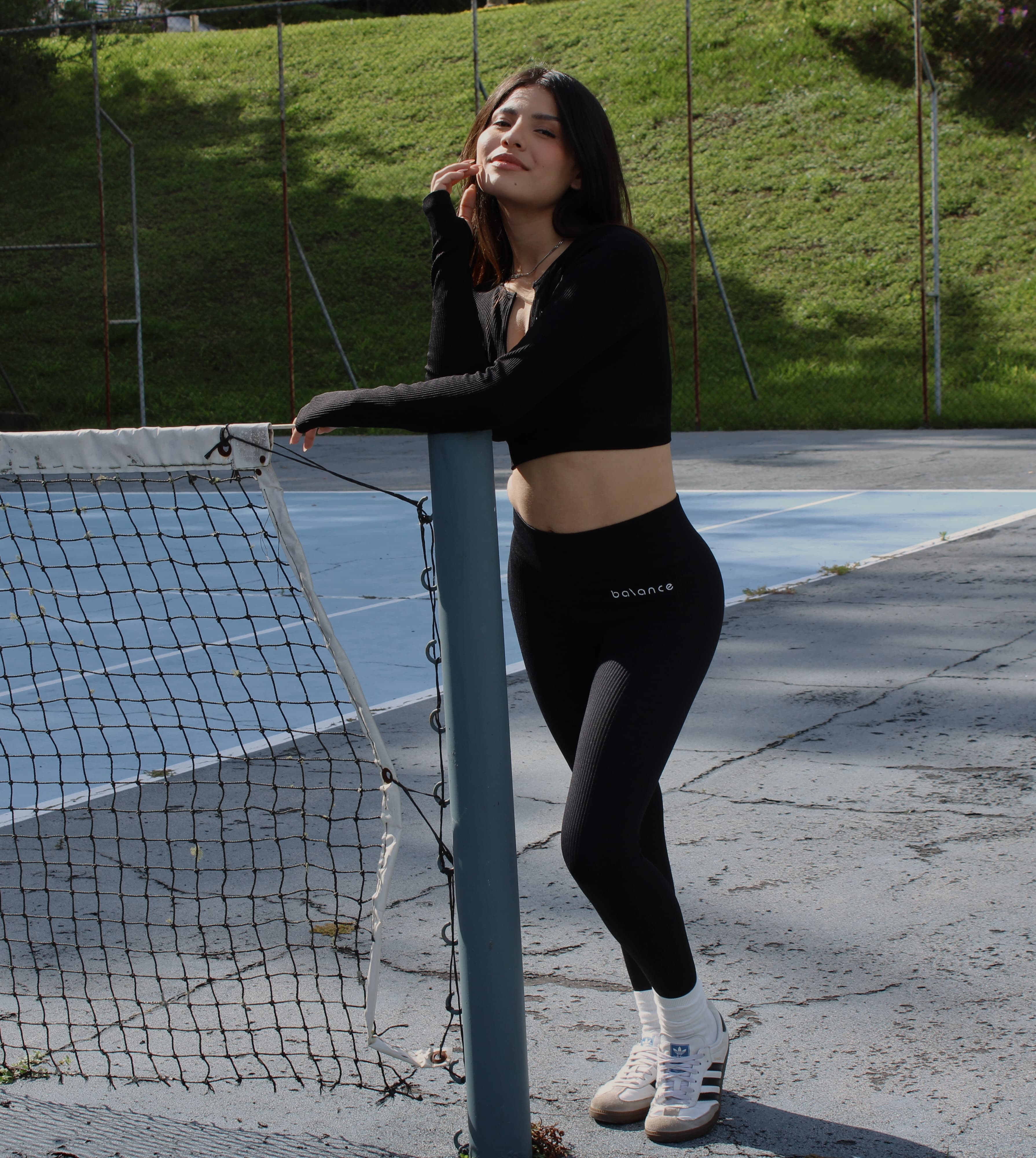 Modelo femenina de pie en una cancha deportiva al aire libre, usando un set de activewear negro con top de manga larga y legging de tiro alto.