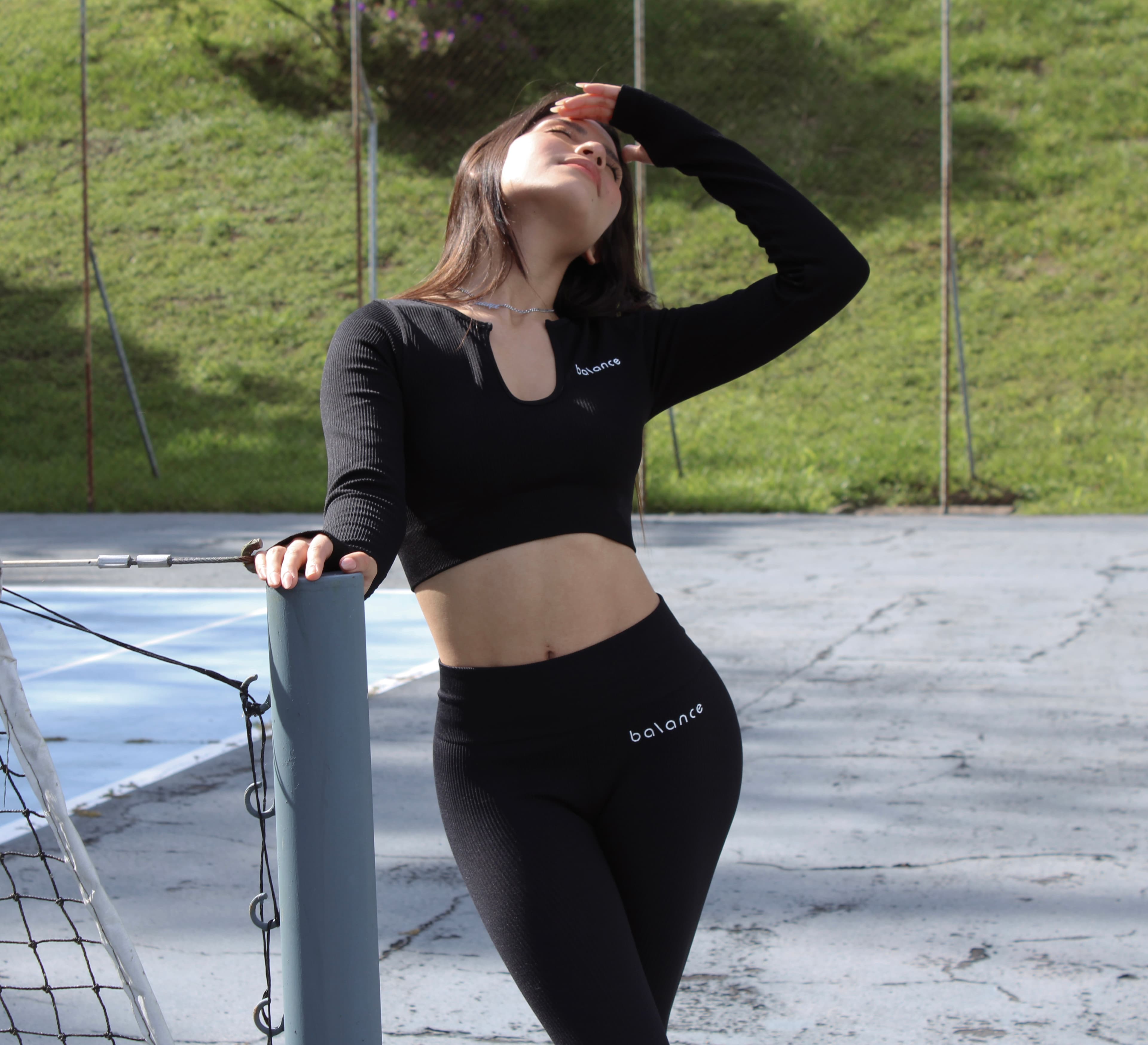 Modelo femenina usando un set de activewear negro con top de manga larga y legging de tiro alto, apoyada en una cancha deportiva al aire libre.
