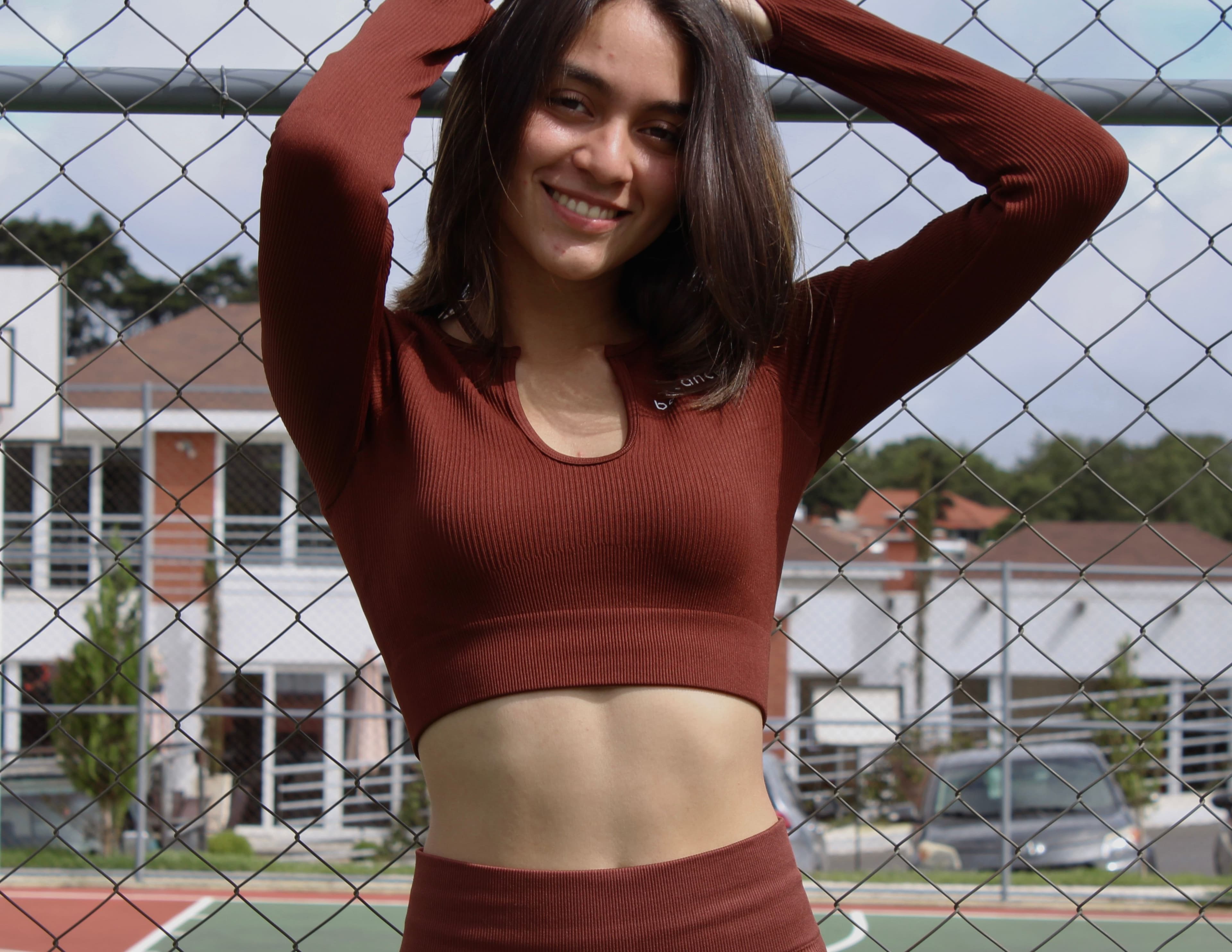 Modelo femenina de frente usando un top de manga larga color marrón, con brazos levantados, en una cancha al aire libre.